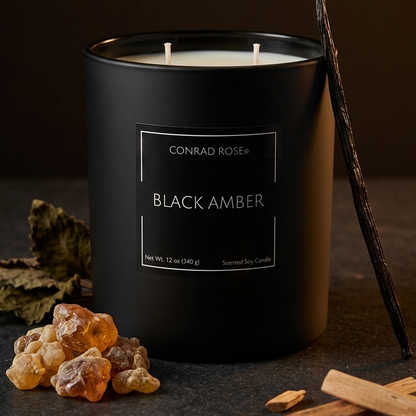 A black matte candle jar labeled 'CONRAD ROSE+ BLACK AMBER' on a dark surface, with cinnamon sticks, amber pieces, and a vanilla bean around it.